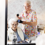Eine Frau frisiert einer anderen Frau die Haare mit einem Fön. Man sieht beide im Spiegel