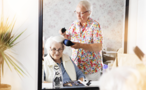 Eine Frau frisiert einer anderen Frau die Haare mit einem Fön. Man sieht beide im Spiegel